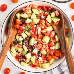 A large bowl of colorful Greek salad, filled with chopped cucumbers, cherry tomatoes, red bell peppers, kalamata olives, red onion, crumbled feta, and herbs. Two wooden salad servers rest on the salad.