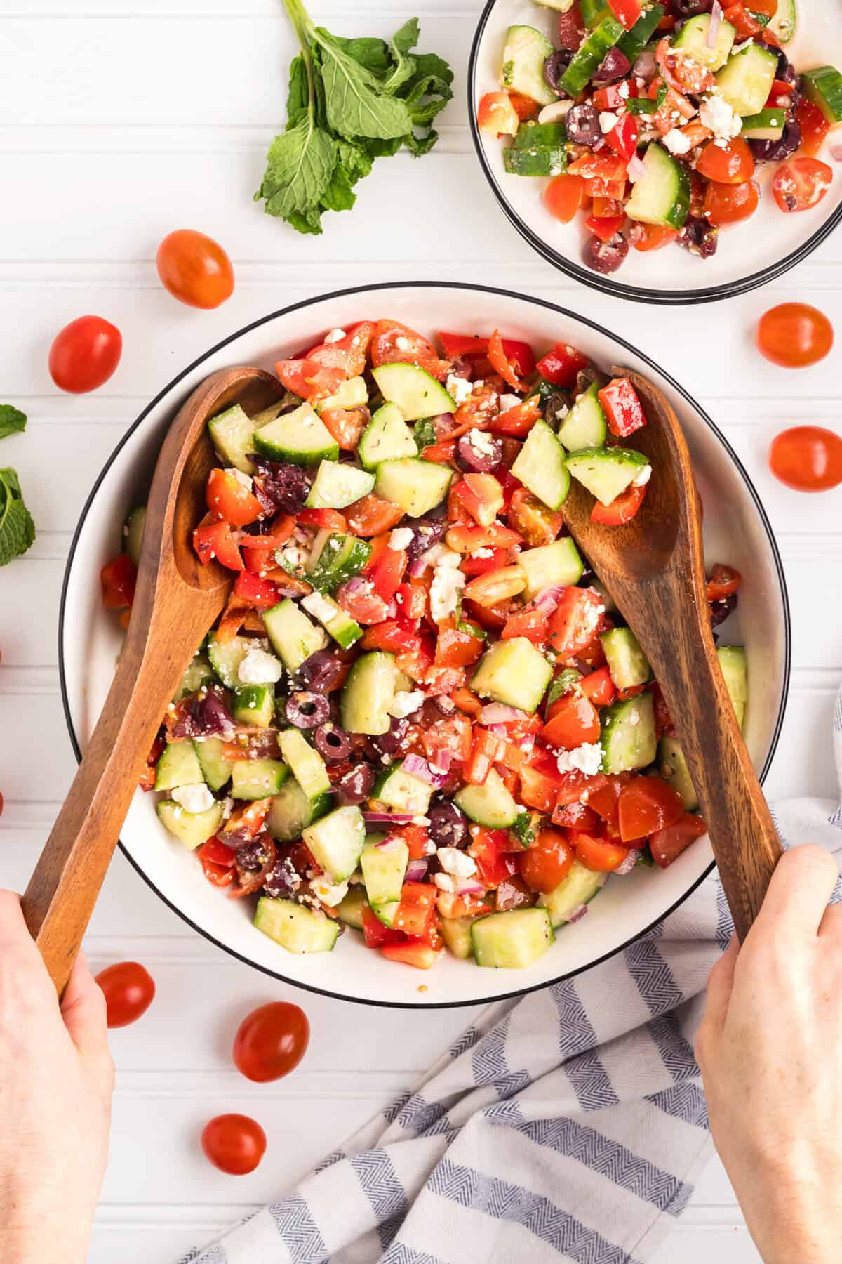 A large bowl of freshly prepared Greek salad with hands holding wooden salad servers, mixing the ingredients. The salad consists of chopped cucumbers, cherry tomatoes, red bell peppers, kalamata olives, red onion, crumbled feta, and herbs.