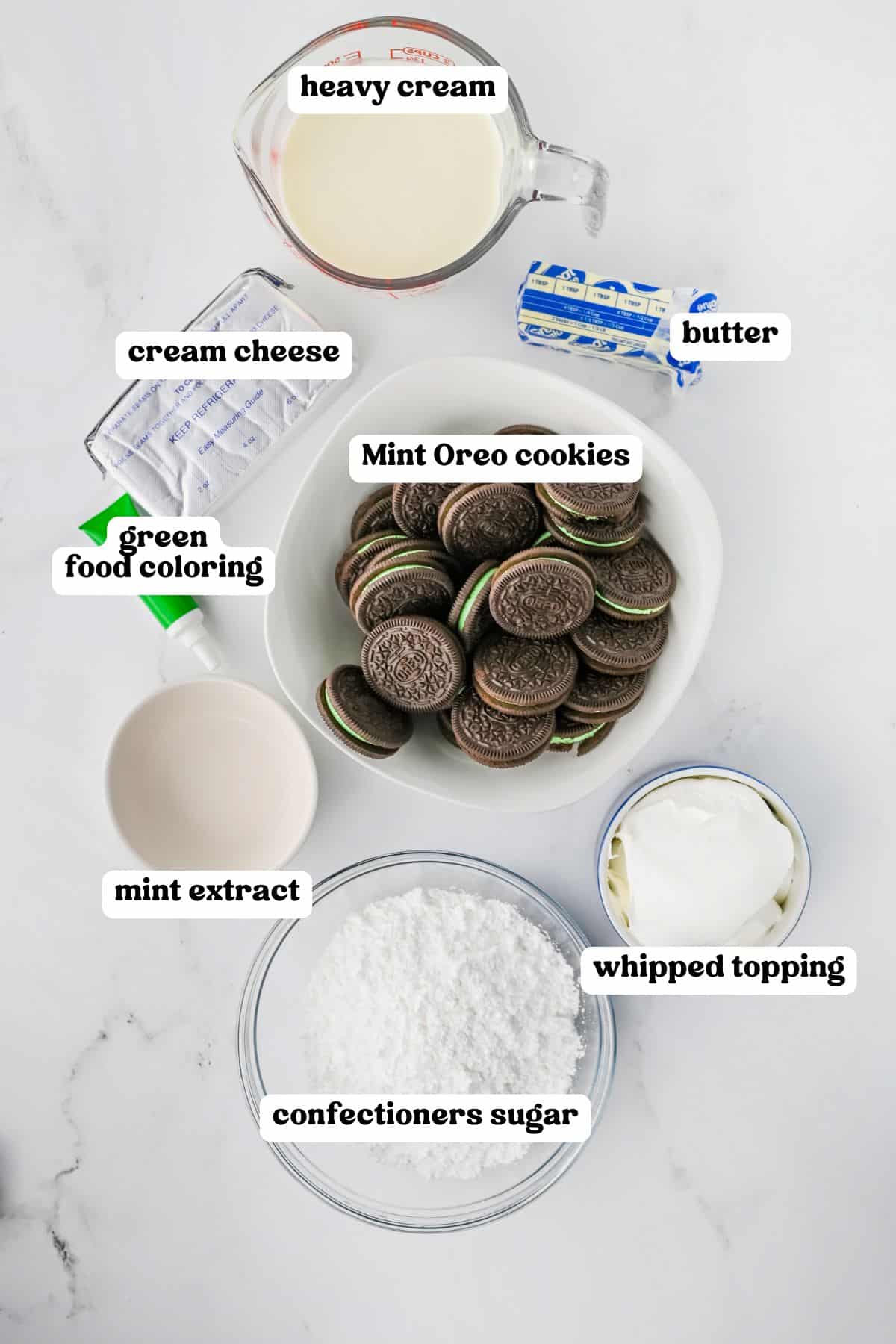 Top-down view of labeled grasshopper mint pie ingredients: heavy cream, cream cheese, butter, green food coloring, Mint Oreo cookies, mint extract, whipped topping, and confectioners sugar.