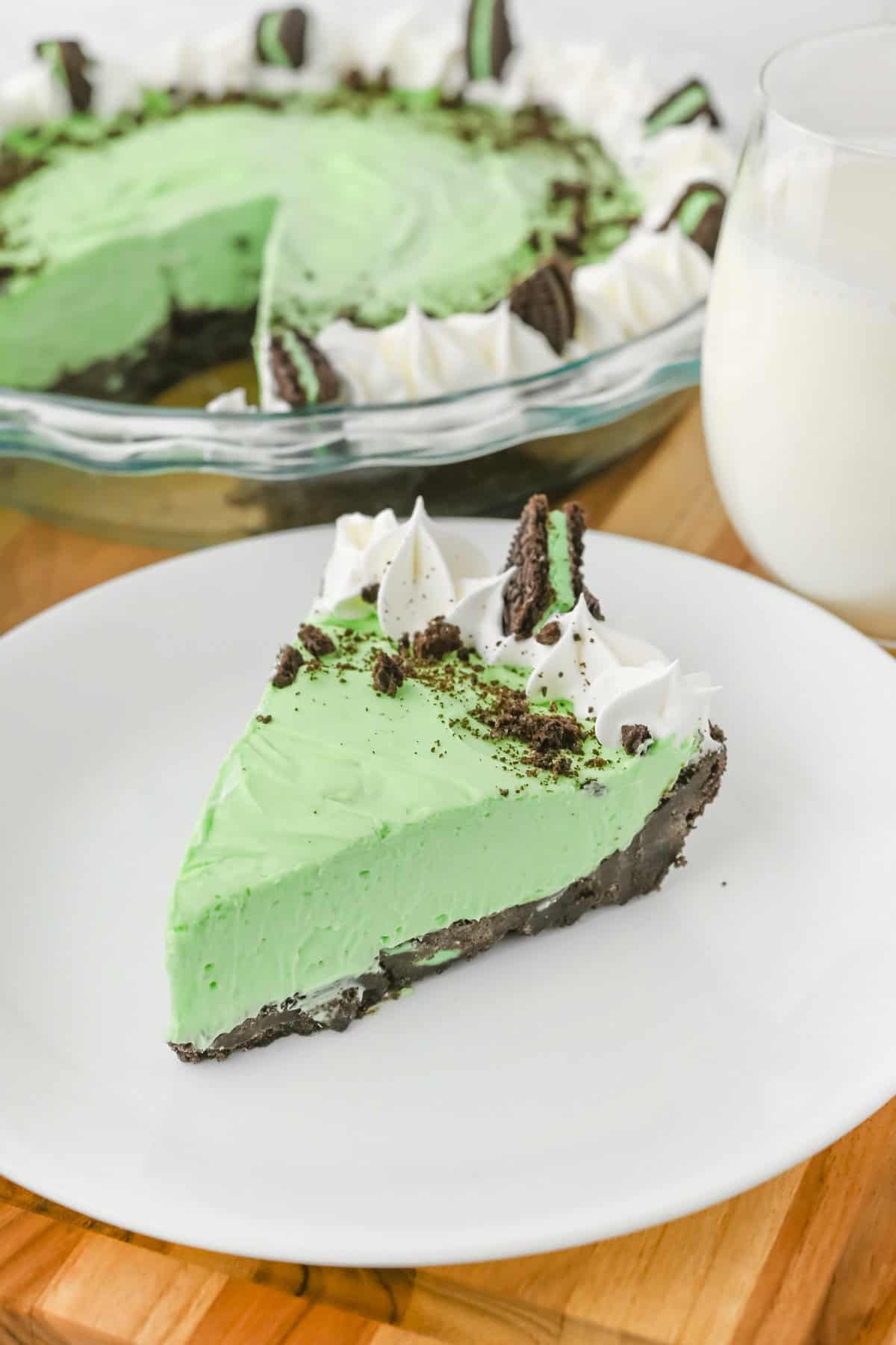A slice of no bake grasshopper pie with smooth green filling, a chocolate cookie crust, topped with whipped cream, crushed cookies, and a Mint Oreo piece, placed on a white plate with the full pie and a glass of milk in the background.