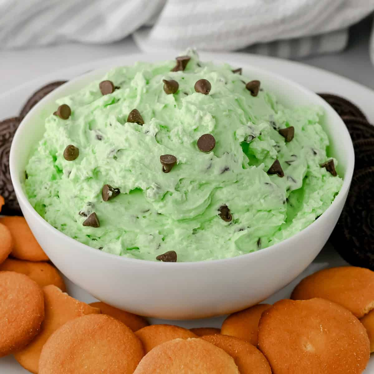 Bowl of mint chocolate chip dip topped with chocolate chips, surrounded by vanilla wafers and chocolate sandwich cookies.