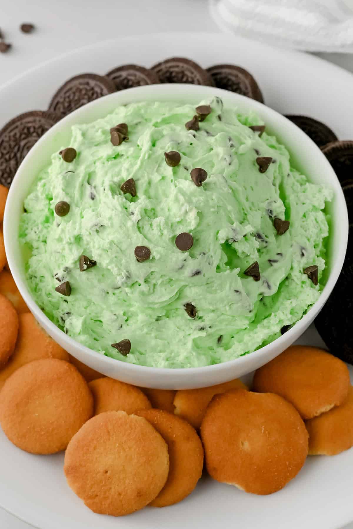 A bowl of creamy green mint chocolate chip dip topped with scattered chocolate chips, served with vanilla wafers and chocolate sandwich cookies for dipping.