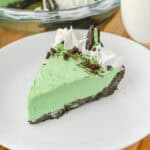 A close-up of a slice of no bake grasshopper pie with green filling, chocolate crust, whipped cream swirls, crushed chocolate cookies, and a Mint Oreo half, served on a white plate.