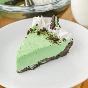 A close-up of a slice of no bake grasshopper pie with green filling, chocolate crust, whipped cream swirls, crushed chocolate cookies, and a Mint Oreo half, served on a white plate.