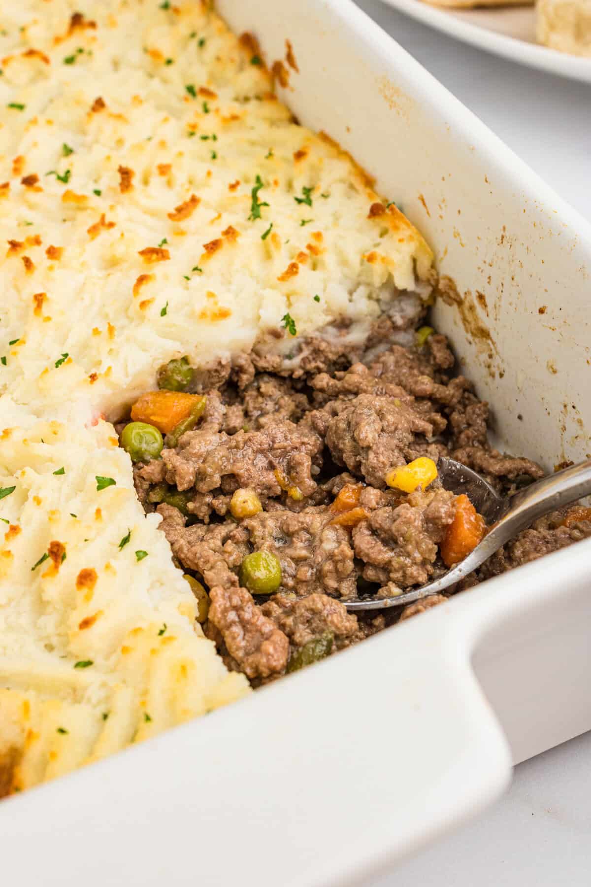 A close-up of a white casserole dish with a portion of cottage pie scooped out, revealing the seasoned ground beef, peas, carrots, corn, and green beans beneath the golden mashed potato topping.