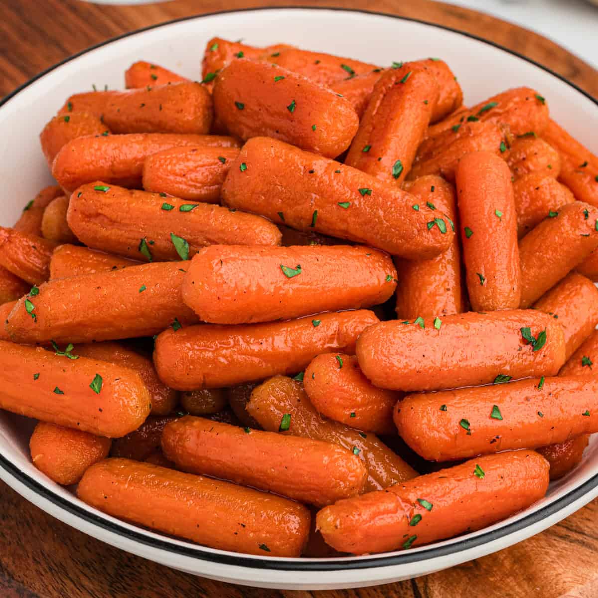 A close-up of a white bowl filled with glossy, brown sugar honey glazed baby carrots, garnished with finely chopped green herbs. The carrots have a slightly caramelized exterior and are arranged in a heaping pile.