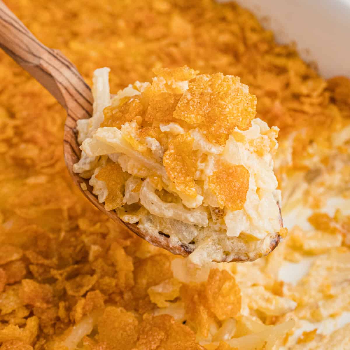 A wooden spoon scooping a portion of cheesy potatoes with hasbrowns with a golden cornflake crust from a casserole dish. The creamy, cheesy texture of the hash brown potatoes contrasts with the crispy topping.
