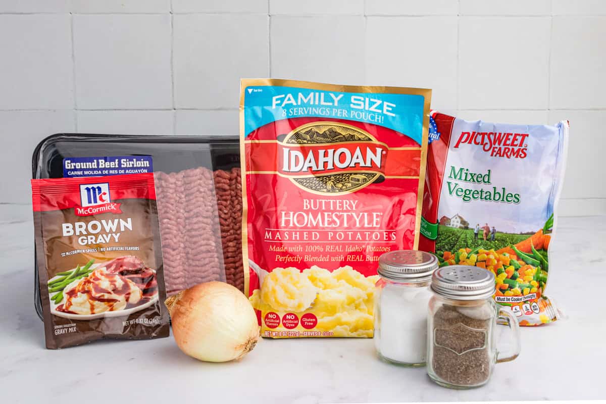 A neatly arranged selection of cottage pie ingredients, featuring a package of ground beef sirloin, McCormick brown gravy mix, Idahoan buttery mashed potatoes, a bag of frozen mixed vegetables, a whole onion, and two glass shakers of salt and pepper.