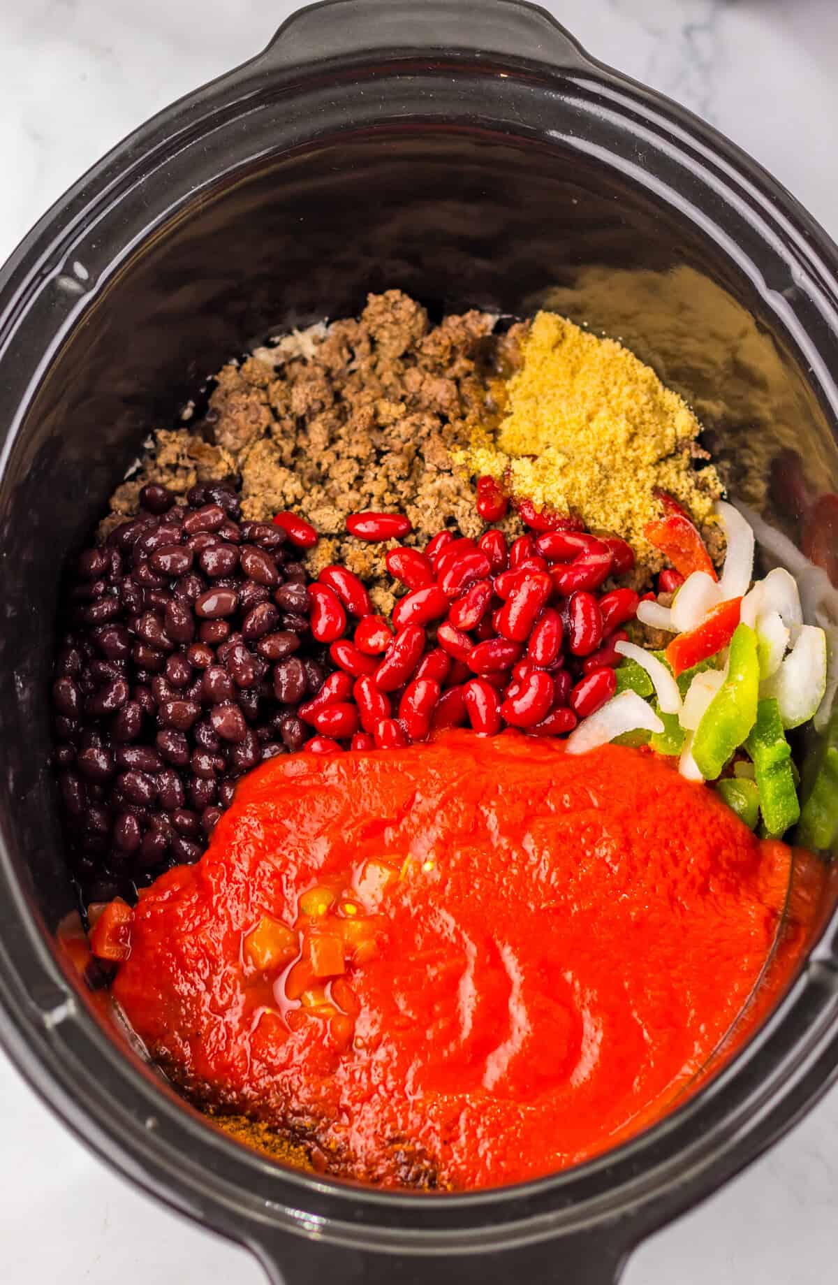 Top-down view of dr pepper chili ingredients in a slow cooker, showing separate piles of black beans, kidney beans, ground beef, tomato sauce, rotel, frozen peppers and onions, light brown sugar, and chili seasoning before mixing or cooking.