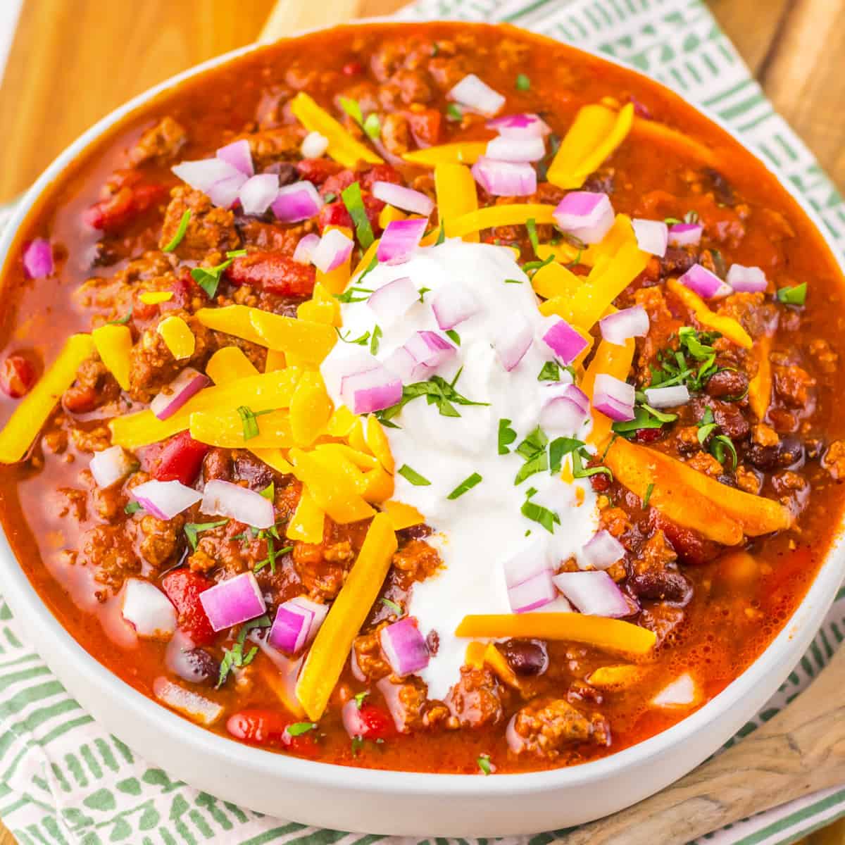 Bowl of Crockpot Dr Pepper Chili with ground beef, beans, and tomatoes in a red sauce, topped with shredded cheese, onions, and a dollop of sour cream.