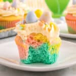 A close-up of a pastel swirl Easter cupcake with a bite taken out, revealing soft layers of blue, pink, and yellow cake. The cupcake is topped with piped buttercream frosting, pastel sprinkles, and three mini chocolate eggs in blue, yellow, and pink.