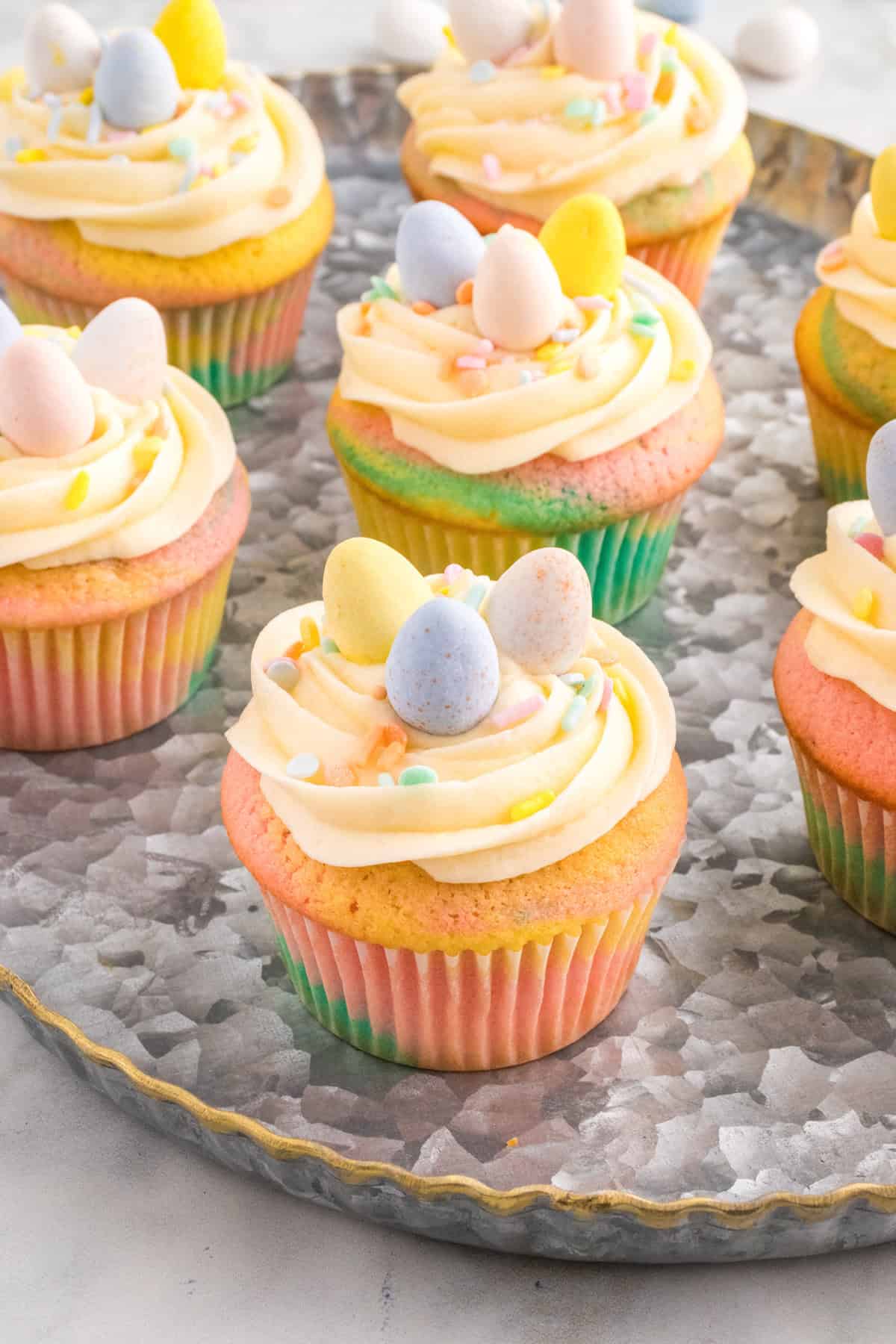 Several pastel swirl Easter cupcakes sit on a round metal tray, each topped with piped buttercream frosting, pastel sprinkles, and mini chocolate eggs.