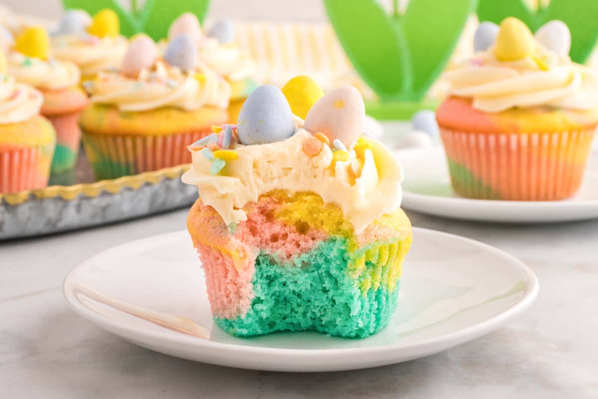 A close-up of a pastel swirl Easter cupcake with a bite taken out, revealing soft layers of blue, pink, and yellow cake. The cupcake is topped with piped buttercream frosting, pastel sprinkles, and three mini chocolate eggs in blue, yellow, and pink.