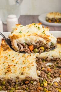 A close-up of a spoonful of cottage pie, showing golden mashed potatoes on top of a beef and vegetable mixture, garnished with chopped parsley. The casserole dish with the remaining pie is visible in the background.