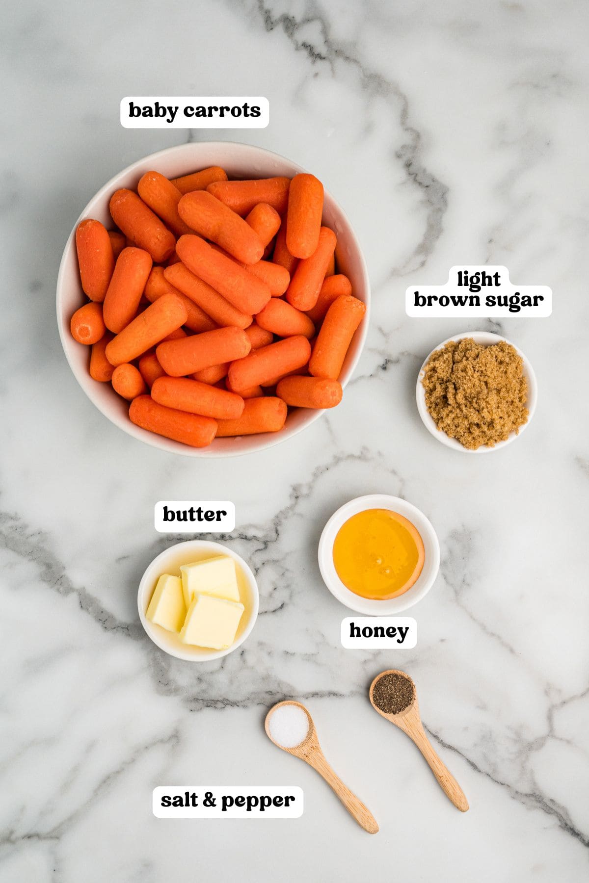 A top-down view of ingredients on a white marble surface, including a bowl of bright orange baby carrots is the largest item. Surrounding it are small dishes containing light brown sugar, cubed butter, golden honey, and spoons filled with salt and pepper.