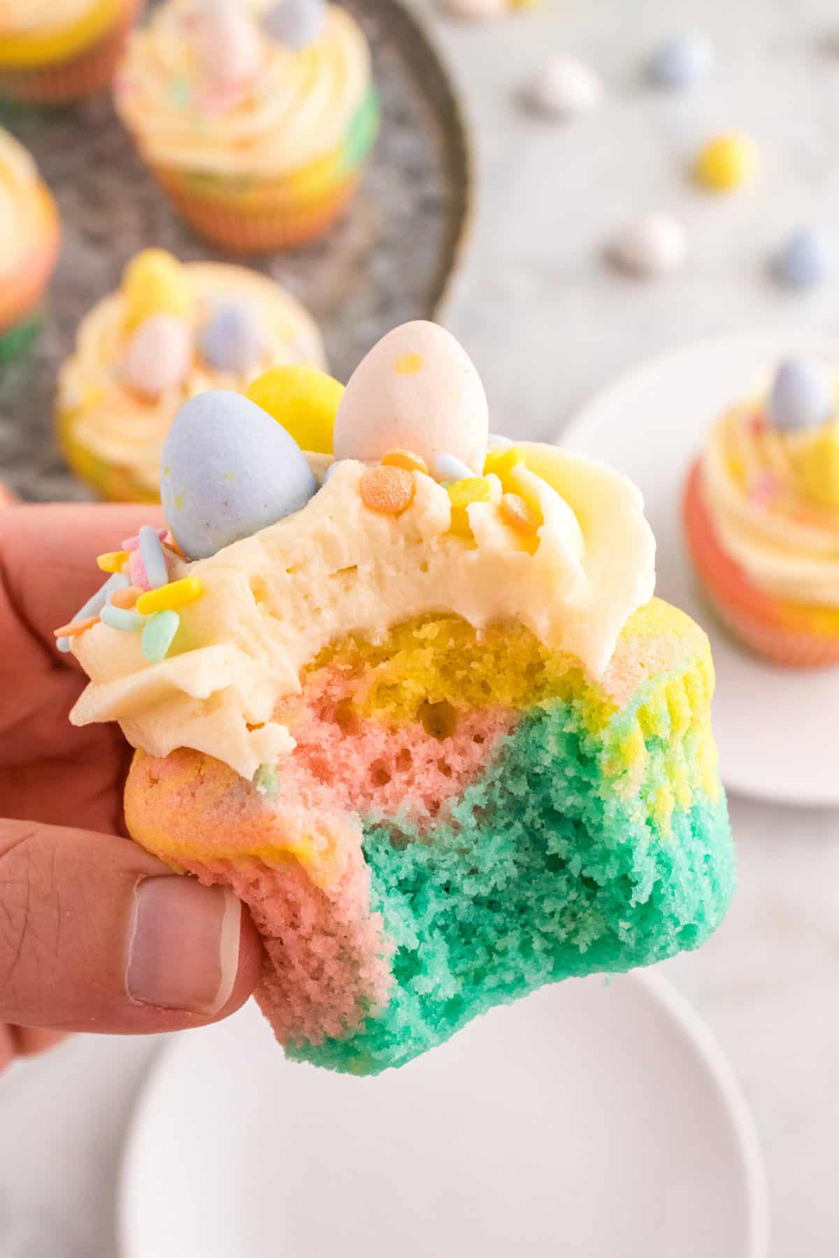 A pastel swirl cupcake held in a hand, showing the colorful blue, pink, and yellow layers inside. The cupcake is decorated with buttercream frosting, pastel sprinkles, and three mini chocolate eggs.