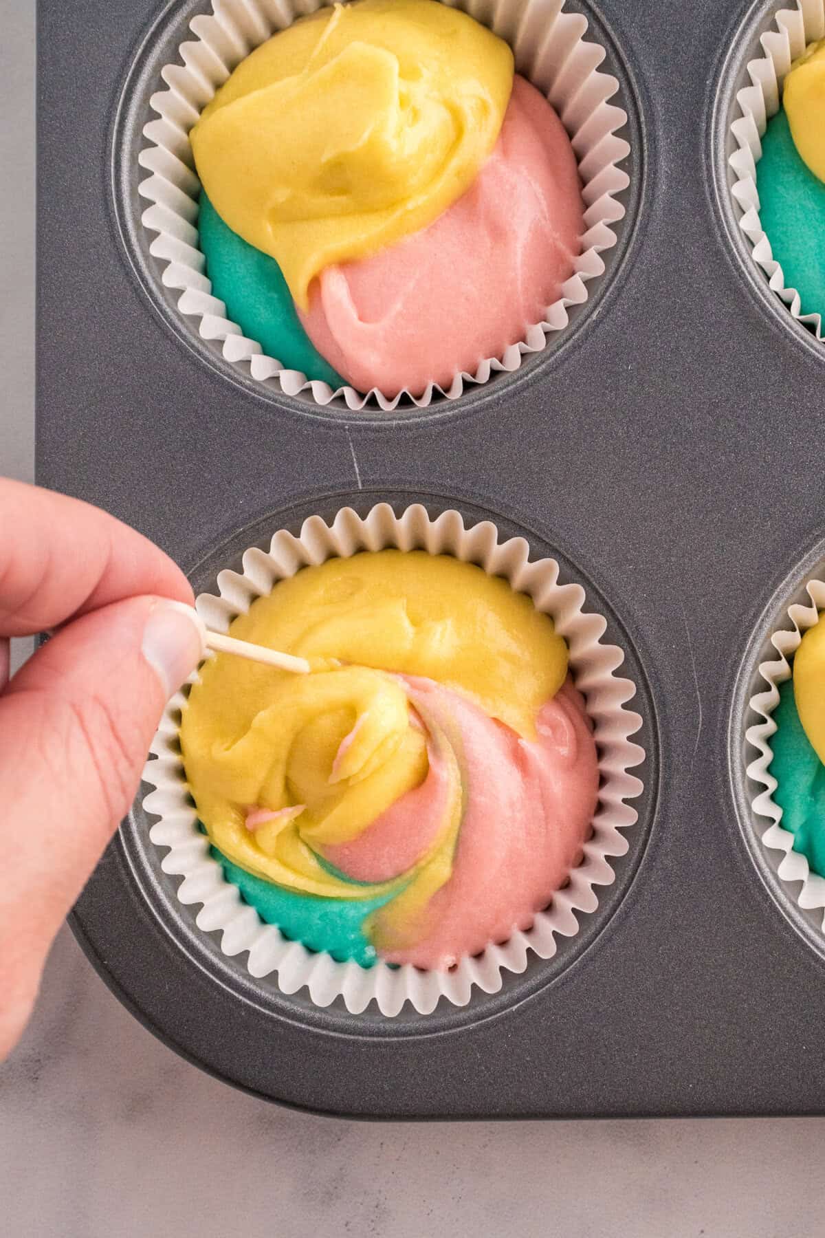 A hand using a toothpick to swirl yellow, pink, and blue cupcake batter inside a paper liner placed in a muffin pan. The colors blend slightly, creating a marbled effect before baking.