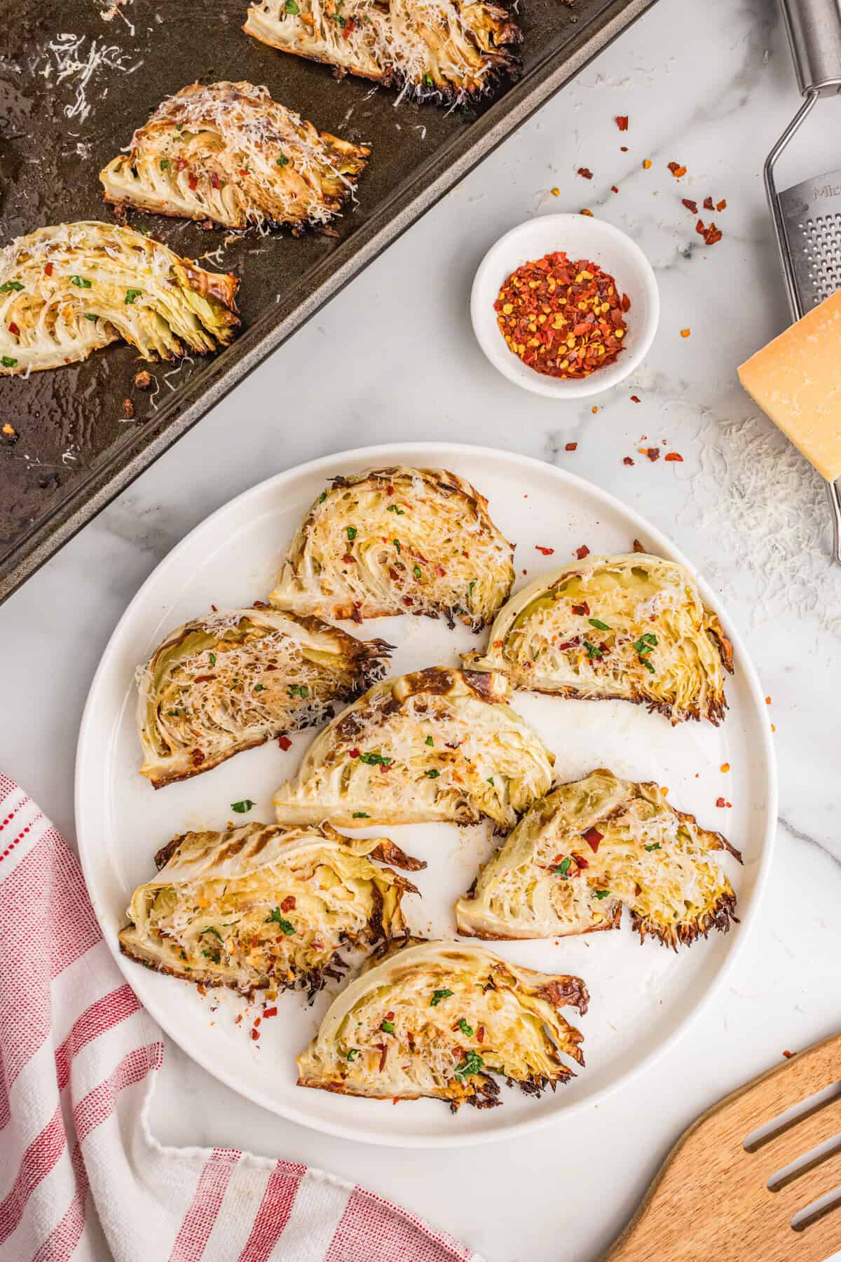 Roasted garlic parmesan cabbage wedges arranged on a white serving plate and baking sheet, topped with shredded parmesan, parsley, and red pepper flakes.
