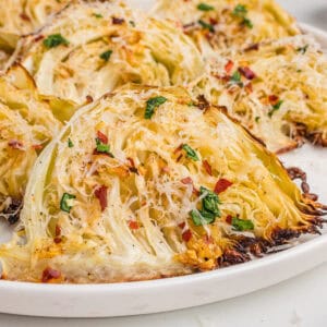 Close-up of roasted garlic parmesan cabbage wedges on a white plate, with golden brown edges, shredded parmesan, parsley, and red pepper flakes.