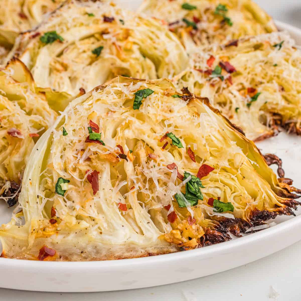 Close-up of roasted garlic parmesan cabbage wedges on a white plate, with golden brown edges, shredded parmesan, parsley, and red pepper flakes.