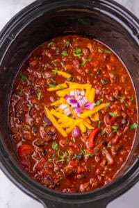 Thick and chunky Dr. Pepper Chili in a slow cooker with visible ground beef, black beans, kidney beans, peppers, and tomatoes. It’s garnished with shredded cheddar cheese, chopped red onions, and fresh herbs.