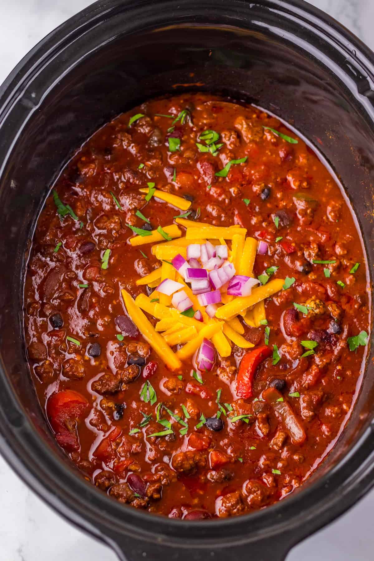 Thick and chunky Dr. Pepper Chili in a slow cooker with visible ground beef, black beans, kidney beans, peppers, and tomatoes. It’s garnished with shredded cheddar cheese, chopped red onions, and fresh herbs.