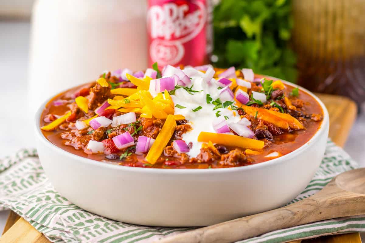 A side view of a white bowl filled with slow cooker dr pepper chili topped with shredded cheddar cheese, chopped red onions, sour cream, and fresh parsley. A can of Dr Pepper and fresh parsley are visible in the blurred background.