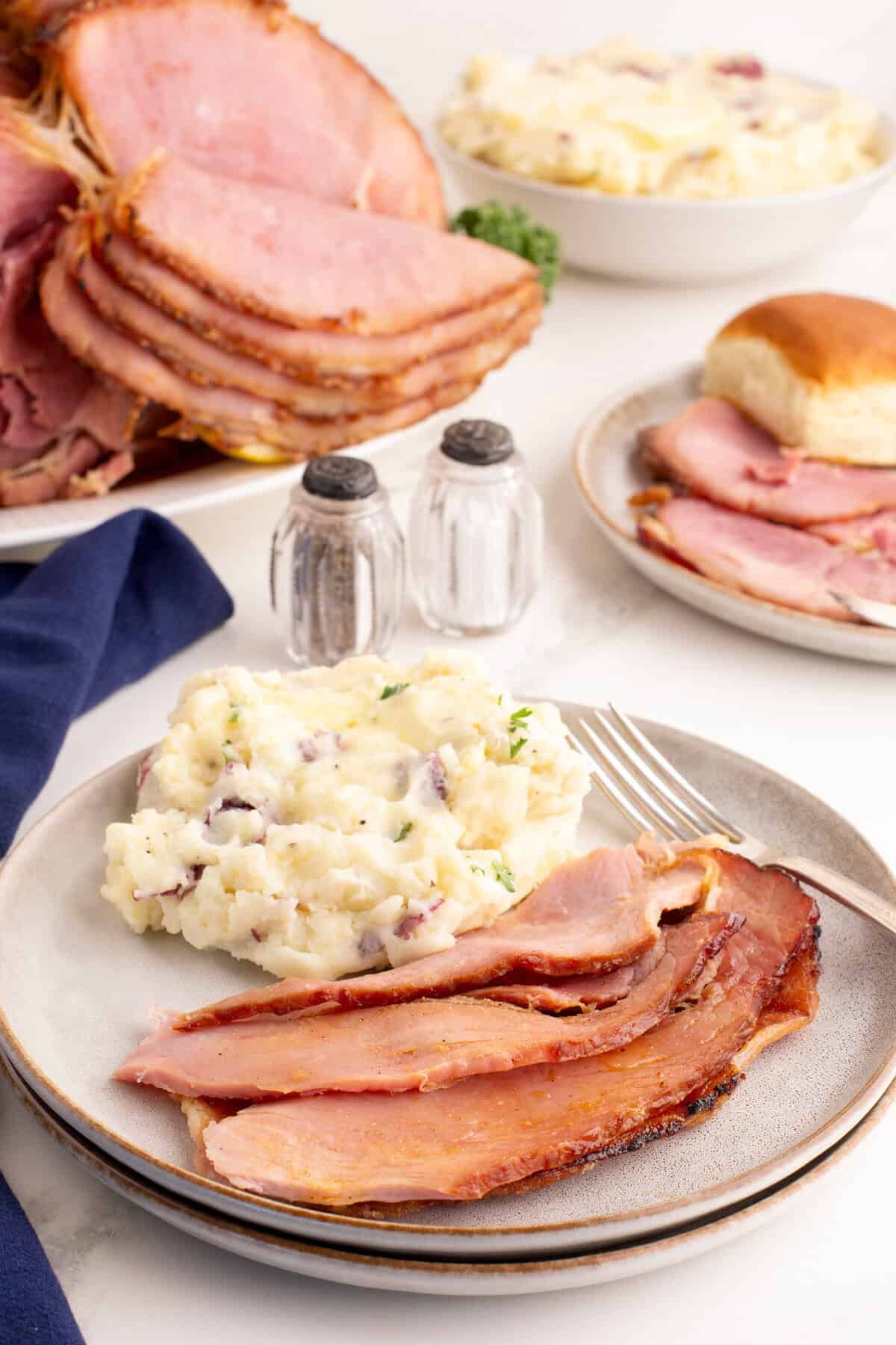Plate of spiral honey ham and mashed potatoes with more spiral cut honey baked ham and sides in the background.