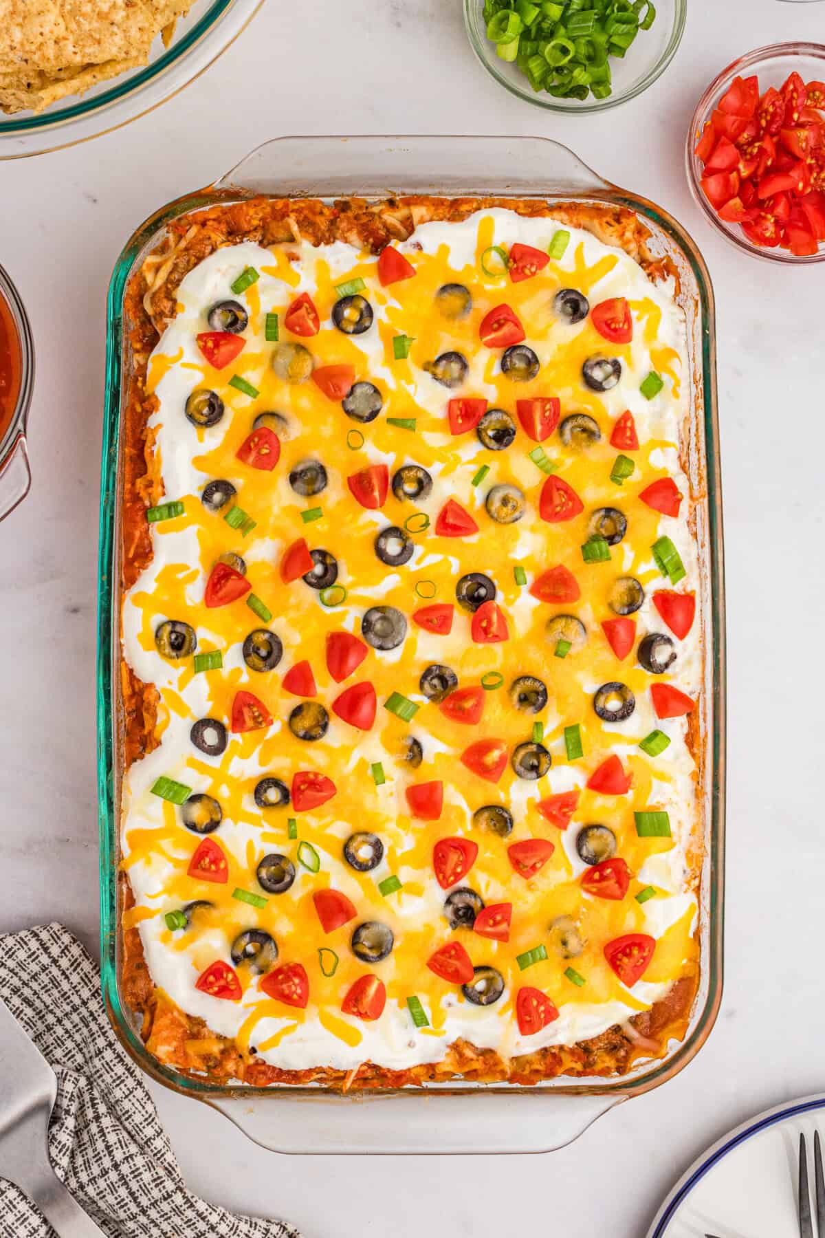 Taco lasagna in a glass 9 x 13 baking dish topped with sour cream, cheese, olives, tomatoes, and green onions.