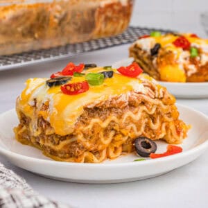 A slice of taco lasagna with noodles on a white plate, topped with melted cheese, olives, tomatoes, and green onions.