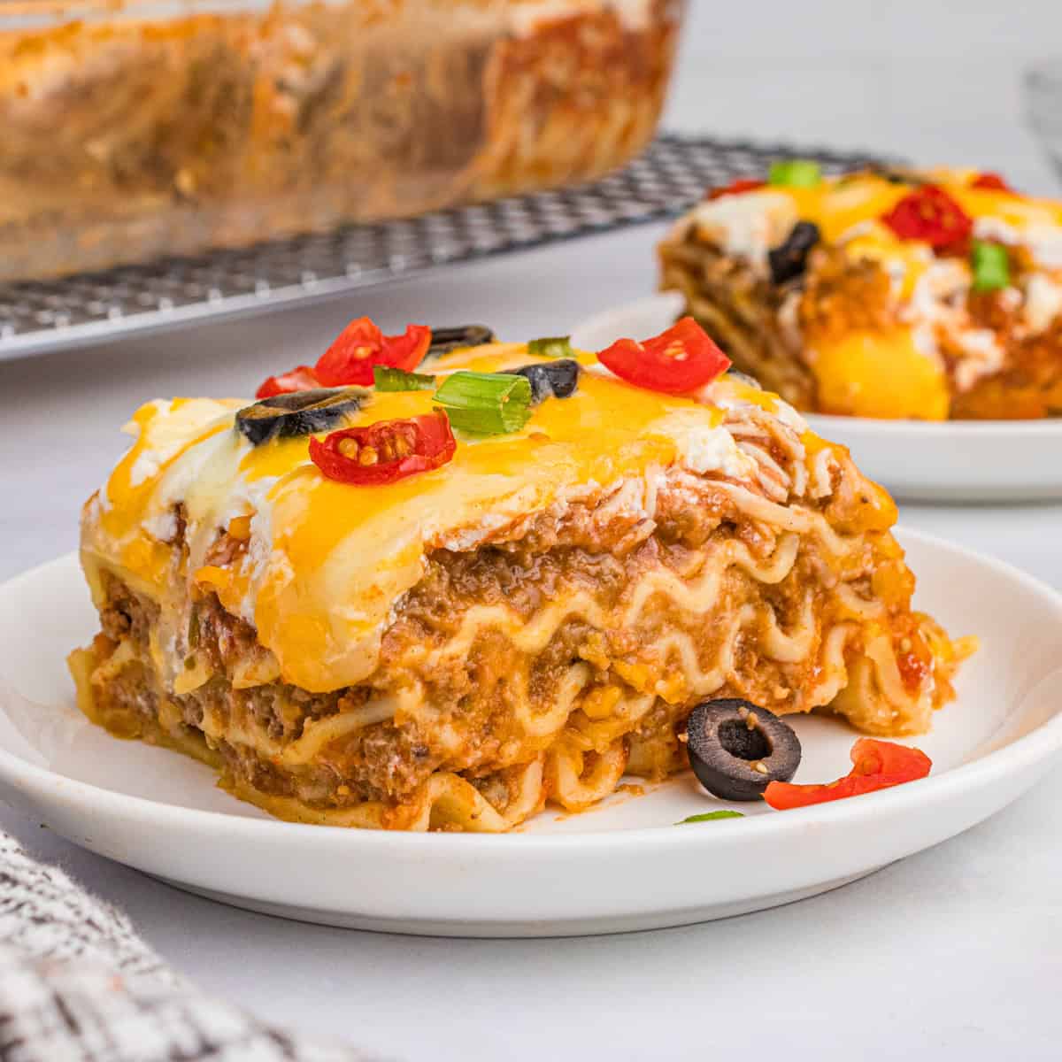 A slice of taco lasagna with noodles on a white plate, topped with melted cheese, olives, tomatoes, and green onions.
