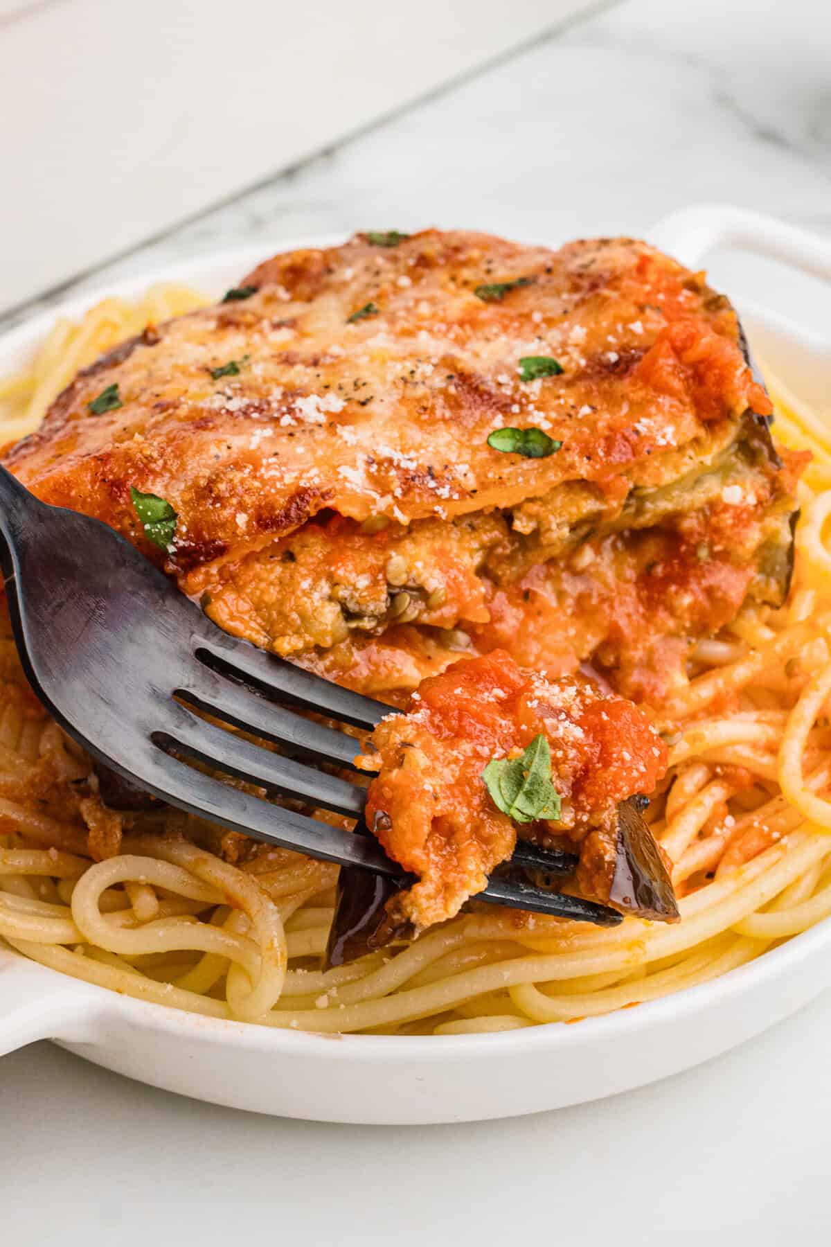 A close-up of a serving of eggplant Parmesan on a bed of spaghetti. A fork is lifting a bite from the saucy, cheesy, and layered eggplant with visible herbs and melted cheese on top.
