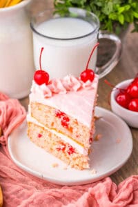 A slice of cherry chip cake with two layers of light pink cake dotted with maraschino cherry bits, filled and topped with pink frosting and decorated with swirls of frosting and whole cherries.