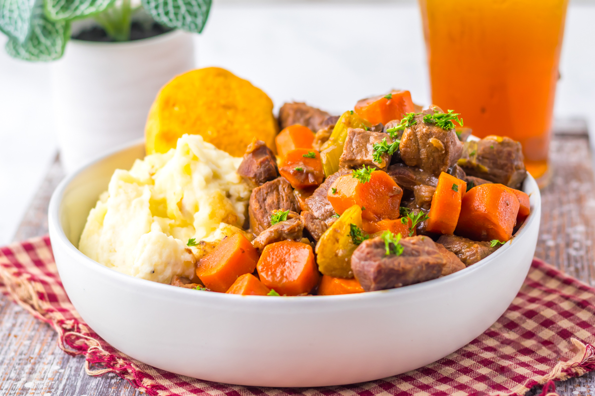Side view of a bowl of crockpot pot roast, featuring stew beef, sliced carrots, celery, mashed potatoes, and a biscuit.