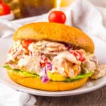 A sandwich filled with creamy Italian grinder chicken salad featuring shredded chicken, pepperoni, salami, provolone, cherry tomatoes, red onions, banana peppers, and lettuce on a golden bun, secured with a toothpick topped with a cherry tomato.