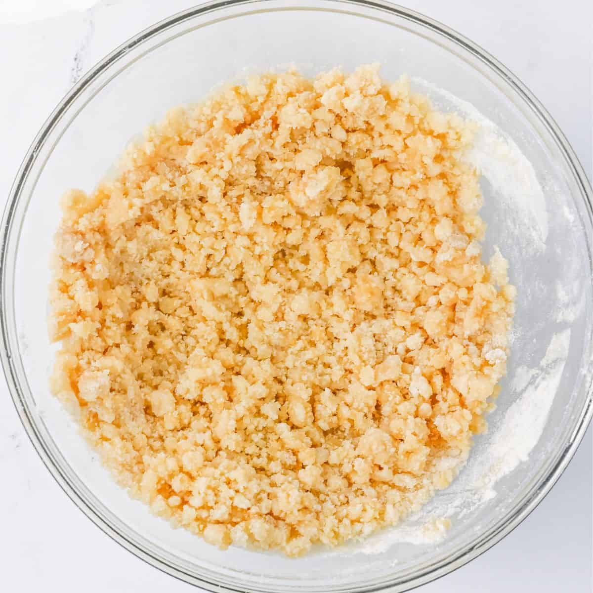 Glass bowl containing a coarse, pale yellow crumb mixture made from flour, butter, and sugar, used for the crumb cake topping.