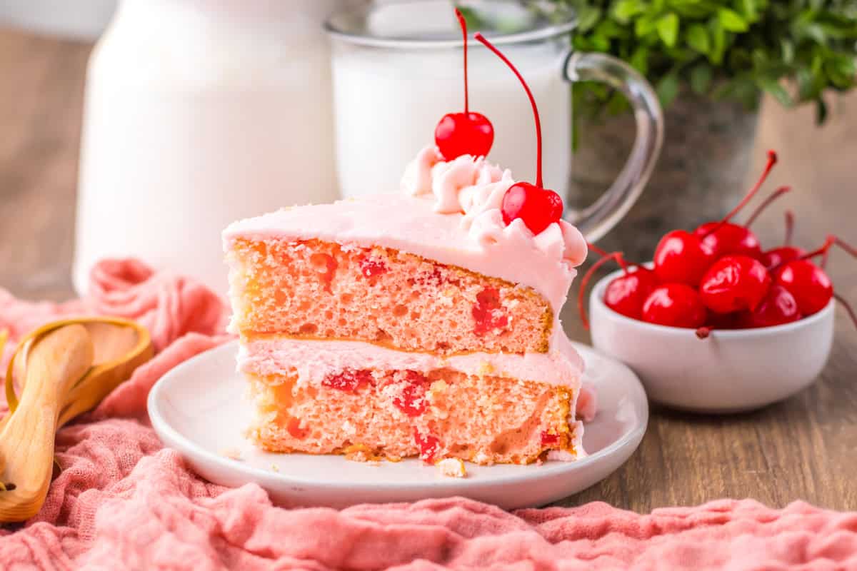 A side view of a cherry chip cake slice showing layers of moist cake with cherry pieces, filled and covered with pink frosting and topped with maraschino cherries.