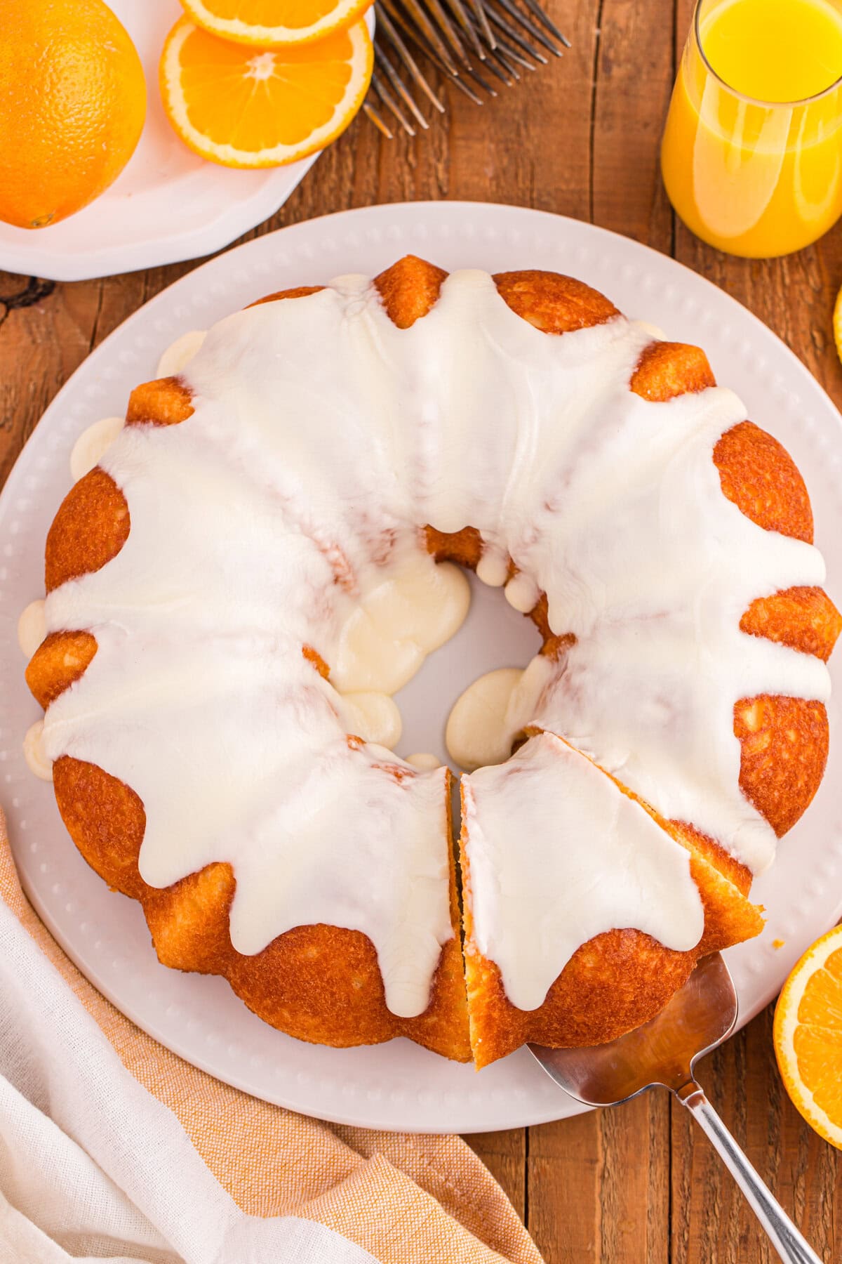 An overhead view of a full orange juice bundt cake topped with white icing. A slice is being removed with a cake server, and orange slices, a glass of juice, and utensils are visible around the plate.