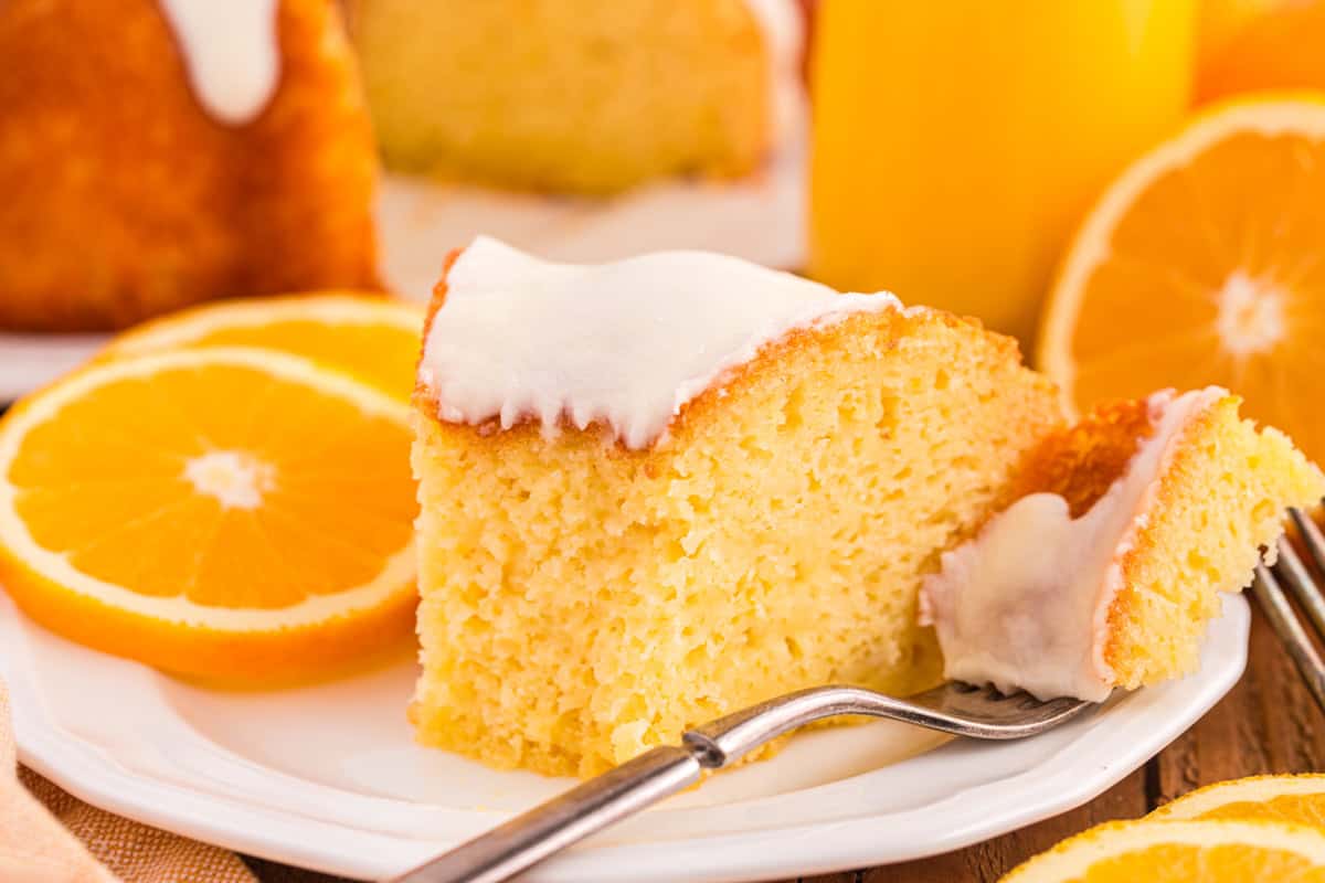 A close-up of a slice of moist orange juice cake topped with a layer of smooth white icing, served on a white plate with fresh orange slices. A fork holds a small bite of the cake.
