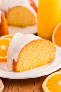A slice of glazed orange juice cake with a smooth white icing glaze sits on a white plate. The background shows halved oranges, a glass of orange juice, and the rest of the cake.