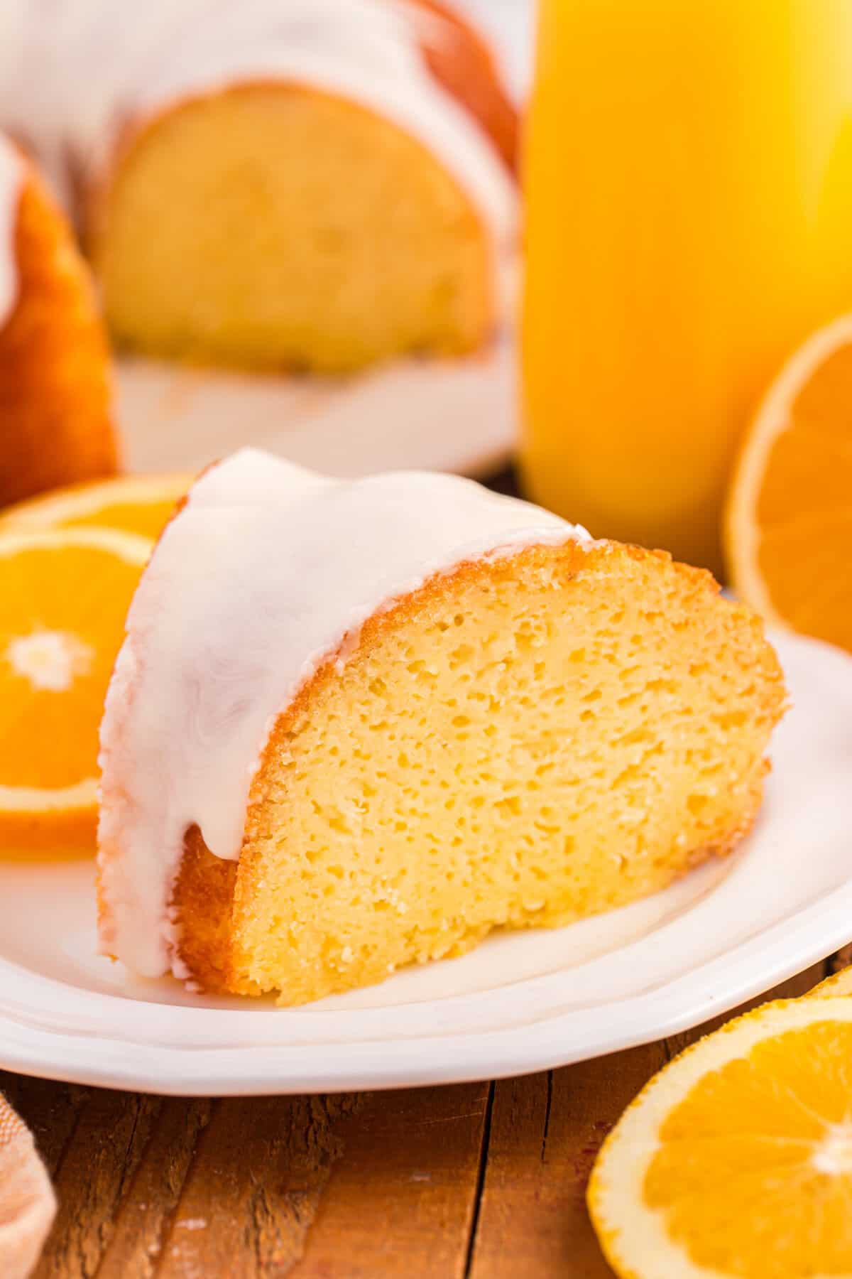 A slice of glazed orange juice cake with a smooth white icing glaze sits on a white plate. The background shows halved oranges, a glass of orange juice, and the rest of the orange bundt cake.