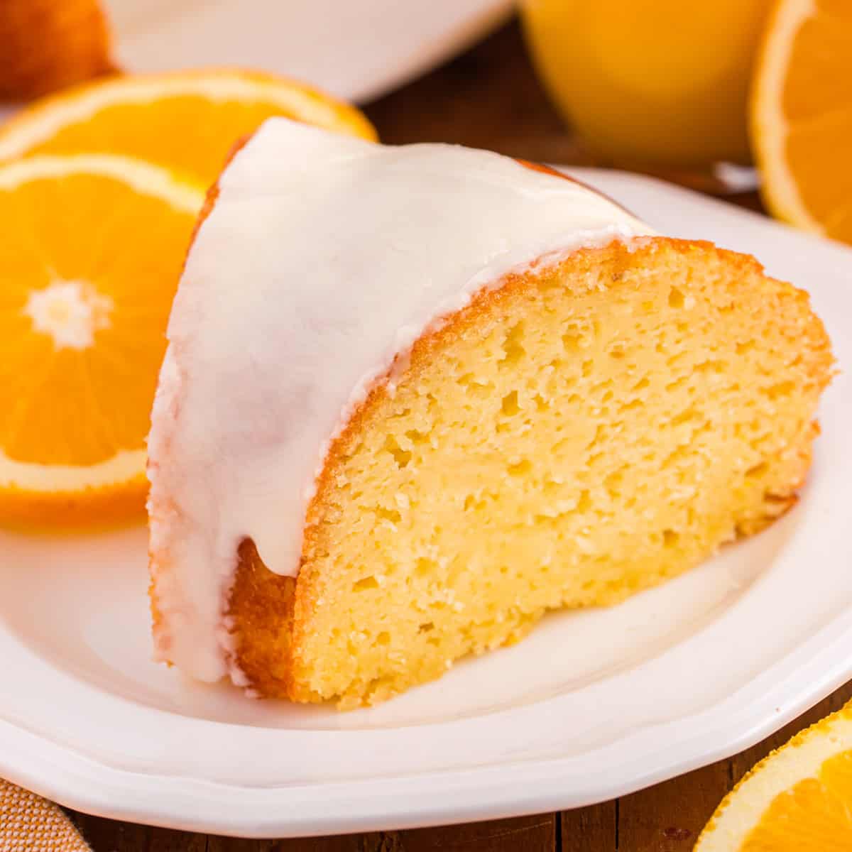 A slice of orange juice cake with a smooth white icing glaze sits on a white plate. The background shows fresh oranges.