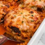 Close-up of a cheesy, golden-brown slice of baked eggplant Parmesan being lifted from a dish, revealing layers of eggplant and sauce with melted cheese on top, garnished with herbs.