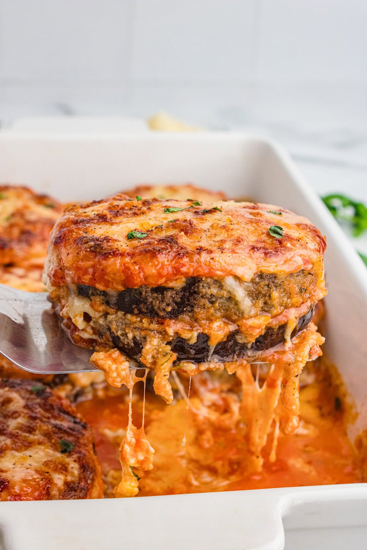 A cheesy slice of oven baked eggplant Parmesan being lifted from the baking dish, with stretchy cheese and visible layers of breaded eggplant, sauce, and cheese.