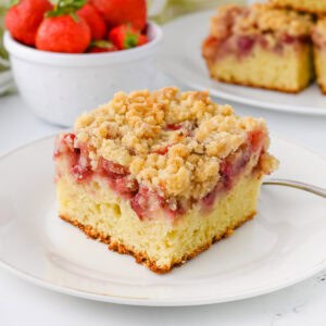 A slice of strawberry crumb cake with a moist vanilla cake base, a layer of baked strawberries, and a golden crumb topping. In the background, there is a bowl of fresh strawberries and additional cake slices.