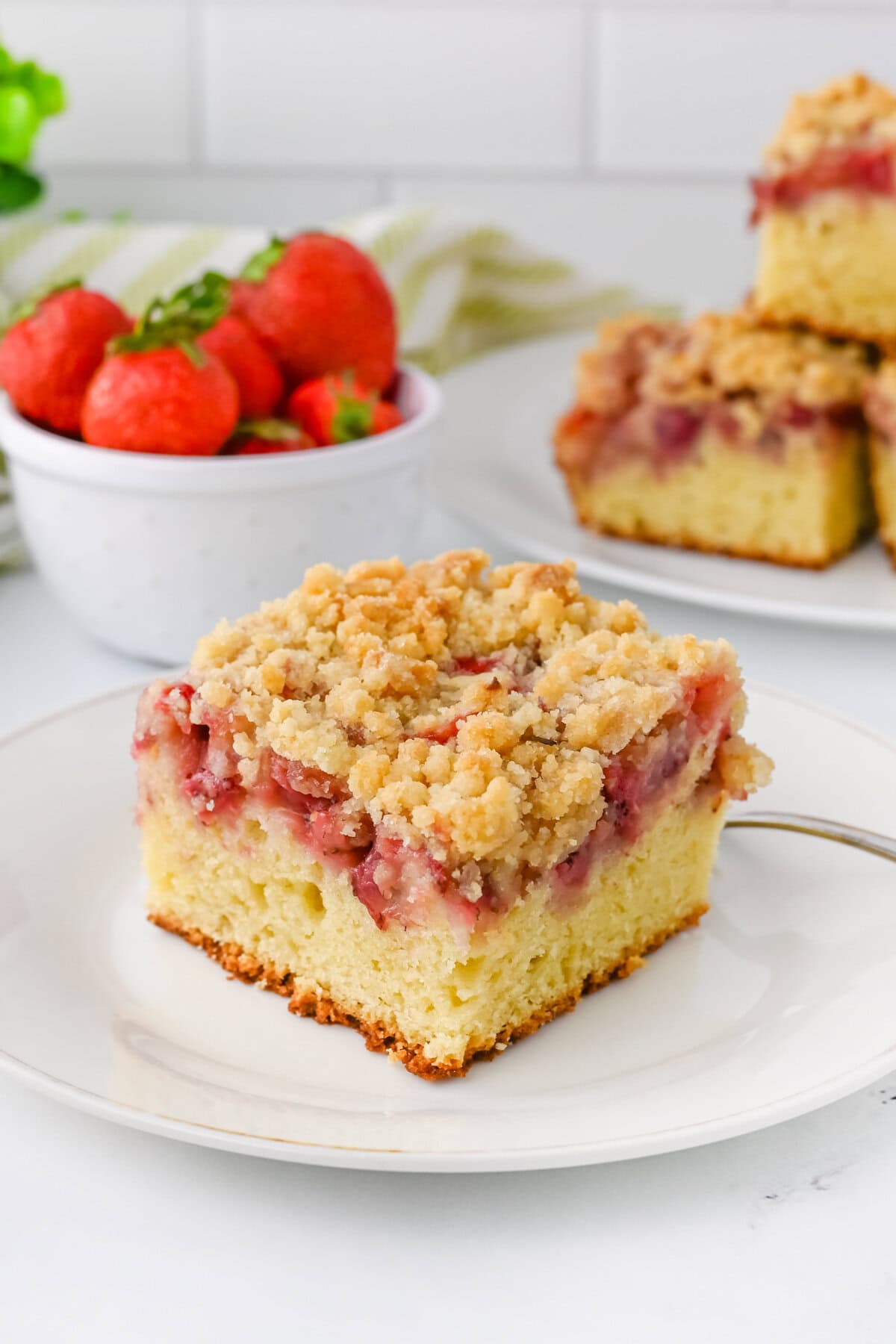 A slice of strawberry crumb cake with a moist vanilla cake base, a layer of baked strawberries, and a golden crumb topping. In the background, there is a bowl of fresh strawberries and additional cake slices.