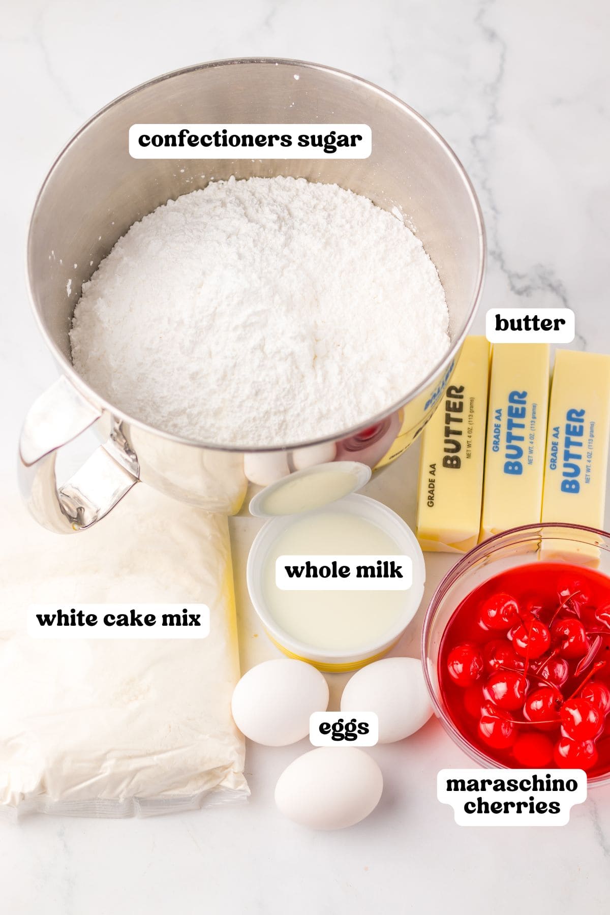 Overhead view of labeled ingredients for maraschino cherry cake, including a metal bowl of confectioners sugar, sticks of butter, a bag of white cake mix, a small bowl of whole milk, three eggs, and a bowl of maraschino cherries in syrup.
