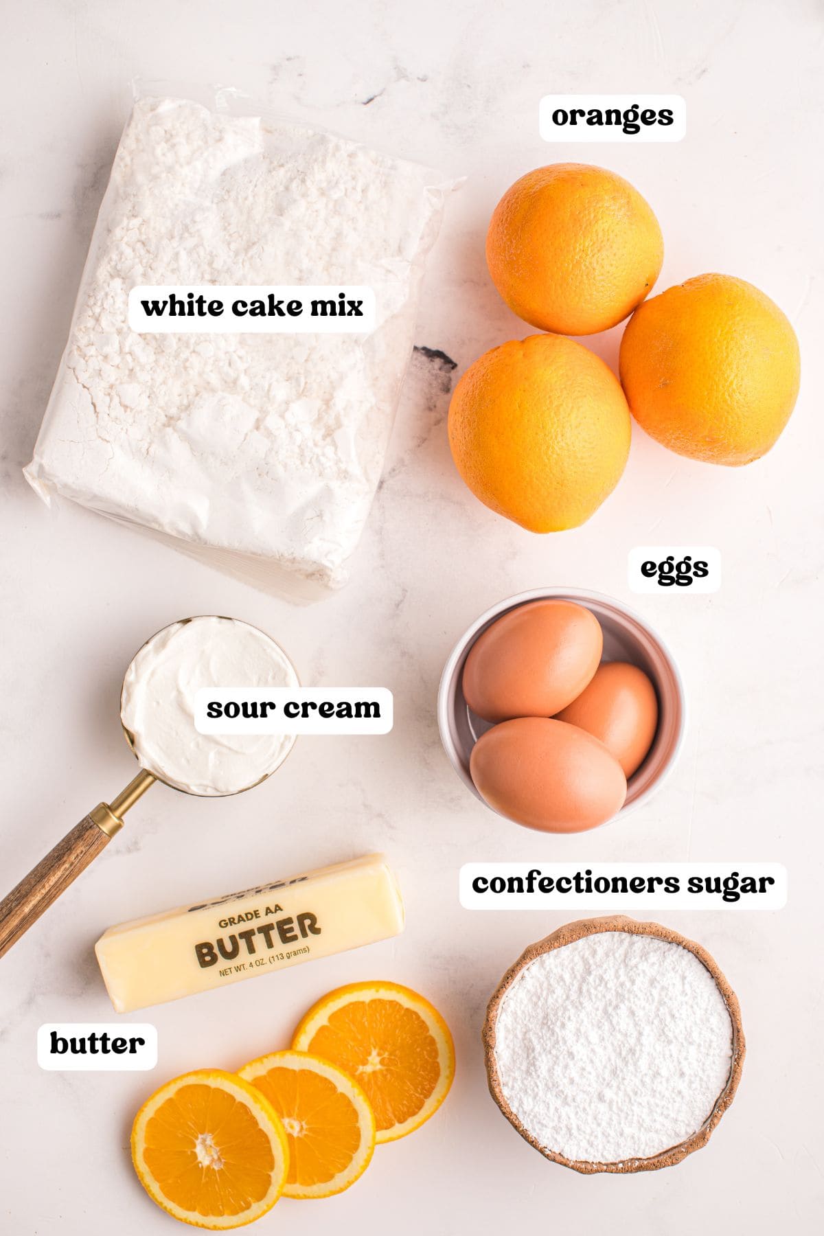 Flat lay of labeled ingredients for orange juice cake on a white surface, including a bag of white cake mix, three oranges, three eggs, confectioners sugar, a stick of butter, and sour cream.
