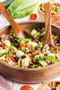 Chopped antipasto salad in a wooden bowl with wooden serving utensils, surrounded by ingredients like romaine, cherry tomatoes, and red onion.