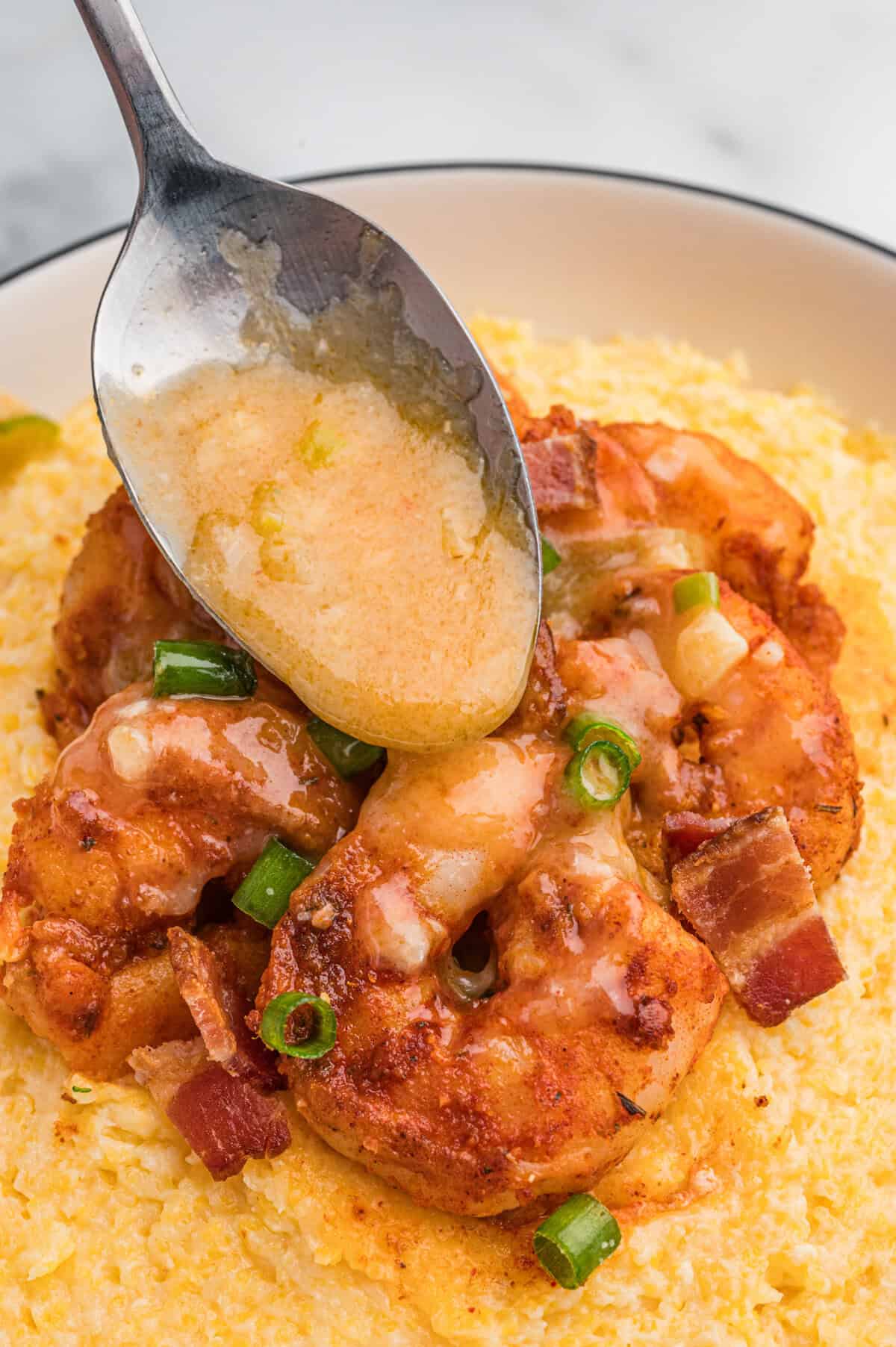 A spoon drizzles buttery sauce over a serving of shrimp on a bed of cheesy grits, garnished with bacon and green onions.